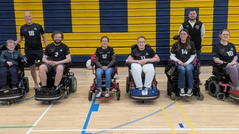 Six people sit in wheelchairs with two men stood behind them. They are on an indoor court with a wooden floor and a blue and yellow wall behind them.