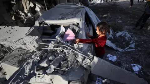 A Palestinian girl inspects a car destroyed by debris following an Israeli strike on a house in Jabalia, northern Gaza (14 May 2025)