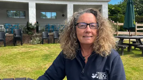 The Image shows Julie Sargent, CEO of Cheltenham Lido, sat on a bench by the Lido's café. She is wearing a blue branded jumper and glasses and smiling into the camera.
