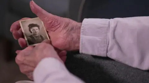 An elderly man's hand is holding a small portrait black and white photo of a young woman with dark hair wearing a shirt. The man holding the photo is wearing a long-sleeved lilac shirt.