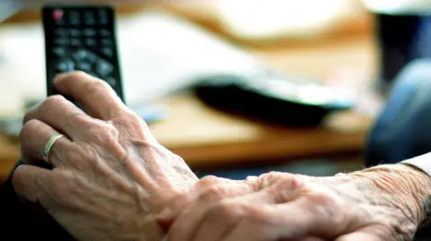 A close-up of a pair of elderly hands, a gold wedding ring can be seen on the left hand holding a television remote.