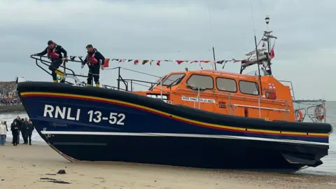 A lifeboat, which has an orange cabin and a navy hull with the lettering "RNLI 13-52" on it. The boat is on a beach and there are two volunteer crew member standing on the deck. People who have come to see the boat are standing nearby on the beach