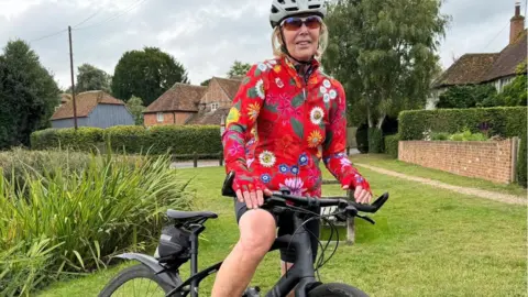 Lin Osbourne saddling her bike and smiling at the camera. She is wearing a grey helmet, brown sunglasses, black cycling shorts and a red flowery cycling jacket.