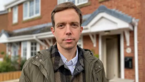 A man with short brown hair is wearing a blue and white striped shirt and a khaki green coat. He is standing outside a house in a street.