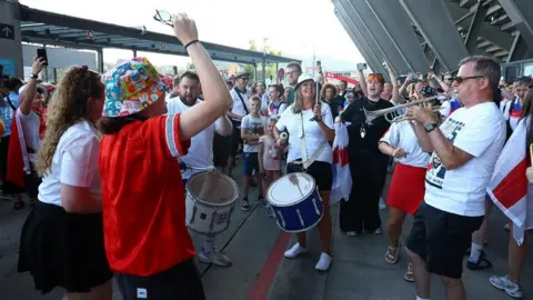 Reuters England fans play musical instruments outside the stadium before the match. The fans, many in hats and wearing white tops, with one in red, are in a circle with the stadium building to one side.