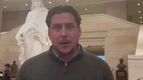 Joe Bennett A man with brown hair stands inside the McCain Institute in Washington. He is wearing a great 1/4 zip jumper and a name badge that reads 'Joe Bennett'. There is a grand marble staircase in the background and a statue. 