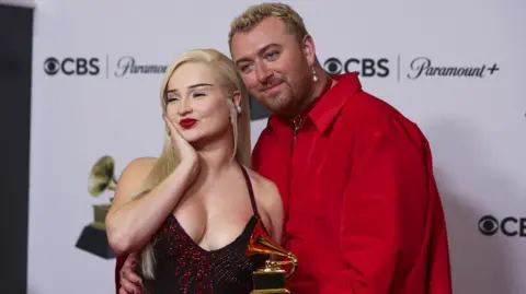 A woman with blonde hair and red lipstick poses with her hand cupping her chin on a red carpet. Standing next to her is a man with short hair and a pearl dangling earring. He wearing a red shirt and has one hand round her waist, with his other holding a small gold trophy.