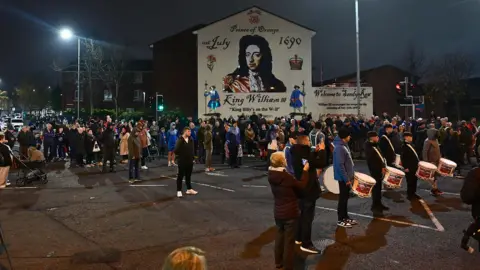 A crowd stand around a mural of William III that's painted on the side of a wall. Some of the protesters are holding drums. It's night.