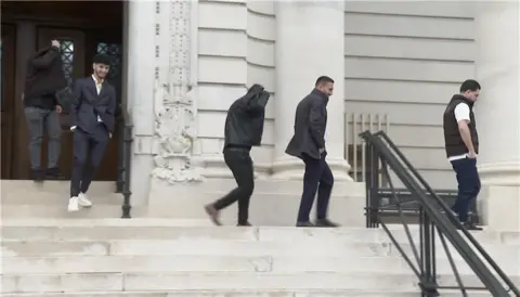 Five men pictured exiting Cardiff Crown Court via the steps. Sardam Ebrahimi can be seen second from left wearing a dark grey two piece suit and he smiles at the camera. He has brown hair and a brown beard. Two others cover their faces with black jackets. The other two defendants, Bave Hamed and Bryar Muradi walk with their hands in their pockets, they both have brown short hair. 