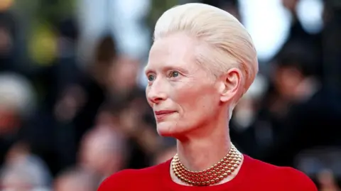 Actress Tilda Swinton, who has short, blonde hair, is dressed in red and wearing a gold necklace. She is smiling and looking to the left.
