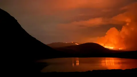 Caleb Emerson Orange smoke rising from the top of a mountain. There is a lake in front of the mountain. 