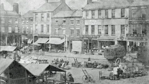 Edwin Williams A grainy black and white image of Preston Market Square showing a row of shops with cloth awnings in the background and stalls, carts and wheeled trolleys in the foreground