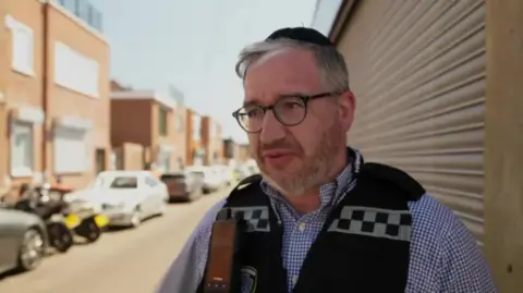 A man white a grey beard and grey hair talks to the camera. He is wearing a black kippah, black glasses, black vest and blue and white chequered shirt. Behind him is a row of houses and a street full of parked cars.