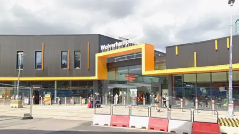 General view of Wolverhampton Railway Station - a large modern rectangular black building with yellow trim. In the foreground there are white and red plastic barriers