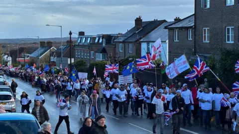 Hundreds of people walking up a hill on one lane of a road. A group at the front of the march are all wearing white t-shirts. Some people are carrying union jacks and East Sussex county flags.