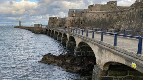 an old stone bridge sits over seawater, alongside a stone castle and leading towards a lighthouse. 