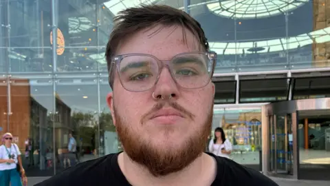 Zoe Dennis/BBC A person with brown hair and a beard looks straight at the camera in a head and shoulders image. They are wearing a black tshirt and grey-framed spectacles and have a septum piercing and a nose ring.