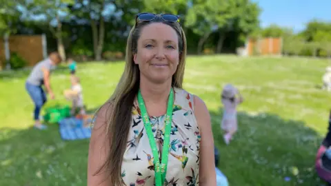 Zara Betts, in a cream dress with birds and wearing a green lanyard. Her hair is long ad is wearing sunglasses on her head. There is grass on the ground behind her. The background is blurred. There is a person behind her and three children. 
There is a fence and trees. 