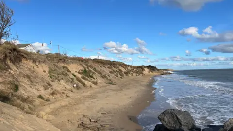 Daniel Hurd A view of a coastline that is being eroded away. A sank bank can be seen reaching several metres above the sea. Patches of grass and hedges are dotted along the bank. 