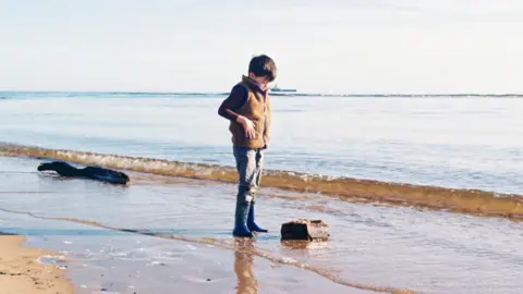 Ian Adamson A Young boy standing at the shoreline, looking down at a rock as gentle waves wash over wet sand on a calm, bright day. He is wearing blue wellies and a tweed bodywarmer.