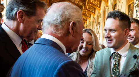 Joseph Holman A close up of the back of King Charles III's head as he talks to Joseph Holman. The king is wearing a blue, pinstriped suit. Holman is wearing a mint-green suit, white shirt and floral tie.