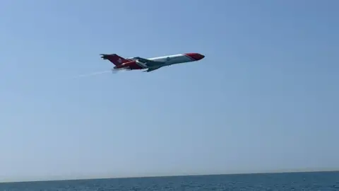 A Boeing 727 tri-jet flying low over the sea on a clear, sunny day. It is painted with red and white livery.