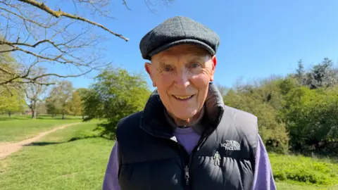 Becki Bowden / BBC A man smiles as he stands on green common land surrounded by trees and hedges. He is wearing a dark flat cap, a black gilet and a purple jumper.