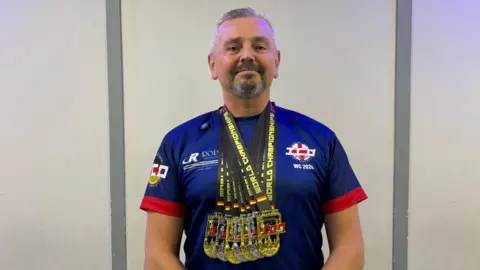 A man with short grey hair and beard wearing a blue and red T shirt and several medals around his neck.