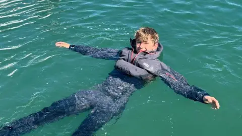 Round Britain eRIB Harry Besley wearing waterproof clothing and an inflatable life vest. He is floating in blue sea with his arms outstretched and smiling