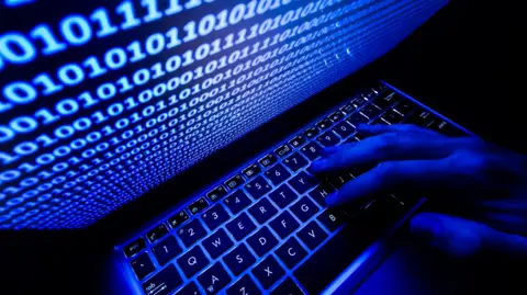 Getty Images An anonymous picture taken in the dark showing a hand on a computer keyboard. A screen above the keyboard is covered with ones and zeros. The picture has a blue tinge, as if from the light of the screen.