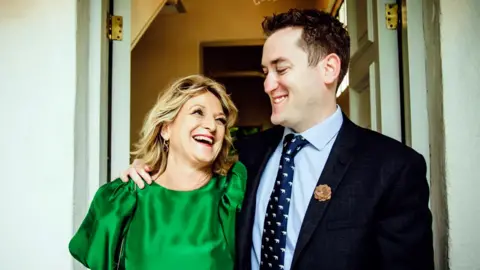 Sam Grice Sam wearing a suit with his arm around his mother in an emerald dress looking at each other and laughing.