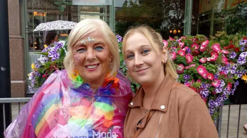 Linda has short blonde hair and is wearing a pink coat, flower chain and poncho. Cassie has long blonde hair in a ponytail and is wearing a brown suede jacket.