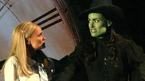Getty Images Kristin Chenoweth as Glinda and Idina Menzel as Elphaba look at each other on stage. Glinda is wearing a white jacket and has blonde hair. Elphaba is painted green and is wearing a black outfit and has black hair. 