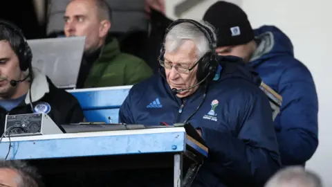Getty Images Football commentator John Helm sat in a commentary position, with a pair of headphones. He is looking down at some notes on his desk.