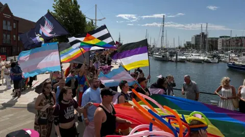 People on the parade hold flags up in the air as they make their way round the marina. Boats can be seen in the background. The water is nearly as blue as the sky.