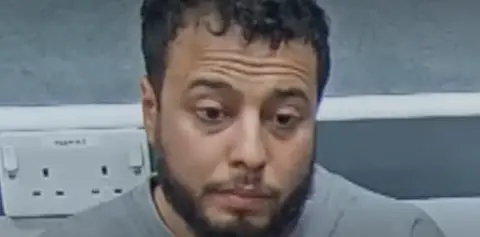 Walid Saadaoui is wearing a grey prison sweatshirt as he is interviewed by police