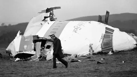 The wrecked nose cone of Pan Am flight 103 lying in a field. A lone policeman is walking infront. The image is black and white