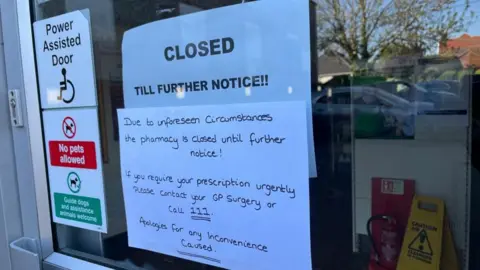 Signs on the glass door at the Wellbeing Pharmacy within the Caister Health Centre site. They read: "Closed till further notice!!!" and "Due to unforeseen circumstances, the pharmacy is closed until further notice! If you require your prescription urgently, please contact your GP surgery or call 111. Apologies for any inconvenience caused"; There are also more permanent signs on the glass saying "Power assisted door"; "No pets allowed; Guide dogs and assistance animals welcome". Reflections of cars parked in the car park, and a wet floor sign and fire extinguisher can be seen on the glass.