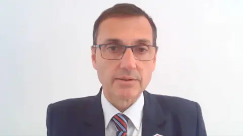 Dr Noble in front of a white background. He is wearing glasses, a dark suit and a tie with red and blue stripes.