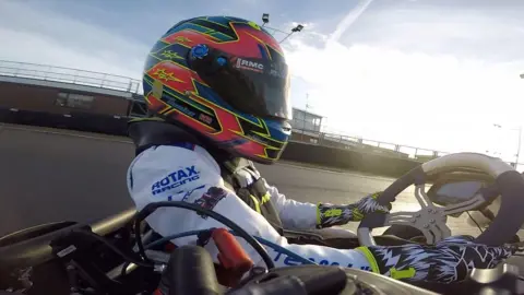 Oliver Cooke Close-up of Josh in a white racing suit and red helmet. He is driving his kart on a race track.