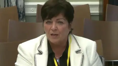 PA Media Devlin has short black hair and is talking in the picture. She is wearing a black shirt and a white blazer. She has a yellow lanyard around her neck.