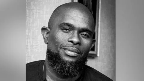 Courtesy Mohammid Walbrook Mohammid Walbrook looks into the camera in this headshot. His head is shaven and he sports a bushy black beard with some grey hairs. 
