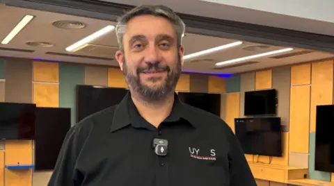 A man with grey hair and a beard is wearing a black short-sleeved shirt. He is looking at the camera and more than half a dozen TV screens mounted on a yellow, green and brown wall behind him are switched off.