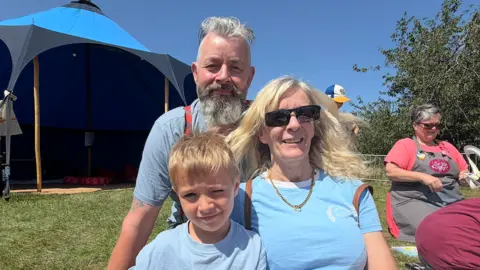 BBC/Jim Scott Pat, on the left and wearing braces around his shirt, sits next to partner Marie, and one of their young family members. They are sitting on the grass and are in front of the blue sensory tent. To the right of them is a woman, wearing pink and grey, and is leading a windmill making workshop.