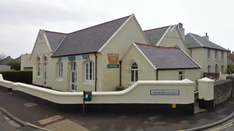 A small former chapel building with a cream exterior and walls surrounding it, which is on the junction of two village roads. 