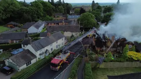 Staffordshire Fire and Rescue Service An aerial image of a rural village with smoke billowing from a thatched cottage on the right. A fire engine is parked in the middle of the road with its ladder extended up to the roof of the cottage. Firefighters can be seen tackling the blaze.