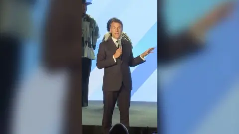 Emmanuel Macron on stage with a mic at Africa Forward summit. He's wearing a dark grey suit and has his hand raised.