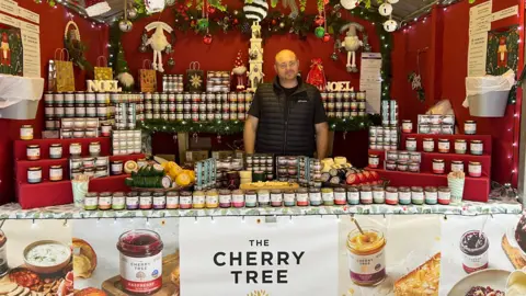 A bald man, wearing a black t-shirt and black body warmer stands in the middle of a cheese and preserves stall. He is smiling and is surrounded by Christmas decorations and jars of preserves and blocks of cheese. 