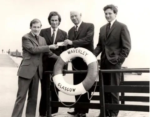 PSPS A black and white image of four men in suits, standing around a Waverley life preserver, one is handing over a pound note to an older man.