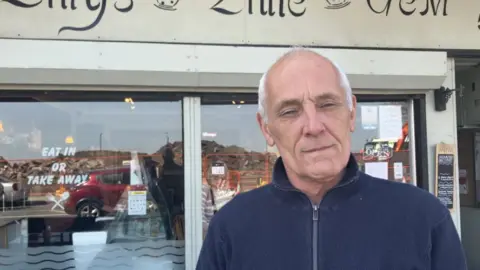 Jonathan Rowbotham - a man with grey hair wearing a dark blue zipper top. He is standing in front of a cafe window.
